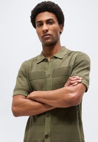 Νεαρός άνδρας με σταυρωμένα χέρια, φοράει μπλούζα πόλο με υφή πλεκτού σε ελιά πράσινο χρώμα και κοντά μανίκια, μπροστά σε απλό φωτεινό φόντο.