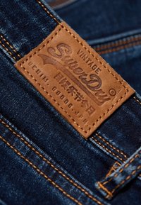 Tummansininen farkkukangas, jossa on erottuva oranssi tikkaus. Sisältää ruskean nahkaleiman, johon on kaiverrettu 'SuperDry' ja lisätietoja.