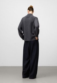 Vrouw met haar in een knot draagt een donkergrijze gebreide trui en brede zwarte broek, met haar handen in de zakken, rechtopstaand met haar rug naar de camera op een grijze vloer.