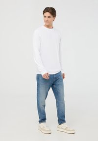 Camicia bianca a maniche lunghe abbinata a jeans azzurri chiari e sneakers color crema. L'outfit presenta una vestibilità rilassata e elementi di design classici.