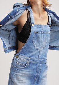 Denimoverall med justerbara axelremmar, ljusblå nyans, framfickor och blekt textur; lager på lager med en denimjacka.