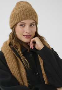 Beige strikket hue med ribbet tekstur, parret med en matchende tørklæde, båret over en sort frakke med store knapper.