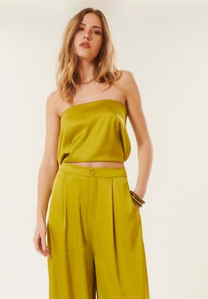 Femme aux longs cheveux portant un top bustier en satin vert olive et un pantalon large taille haute assorti, accessoirisée d’un collier et de bracelets dorés.