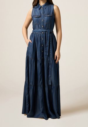Robe longue en denim bleu foncé sans manches avec boutons sur le devant, deux poches poitrine et ceinture nouée à la taille, portée par une femme avec la main dans la poche.