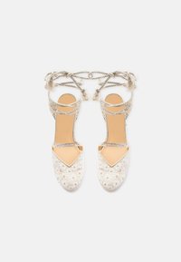 Paire de sandales espadrilles en dentelle beige avec broderies florales et brides tressées à nouer autour de la cheville sur fond blanc.