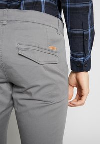 Graue Hose mit einer Gesäßtasche und einem aufgenähten Etikett. Der Stoff wirkt glatt und hat eine leicht matte Textur.