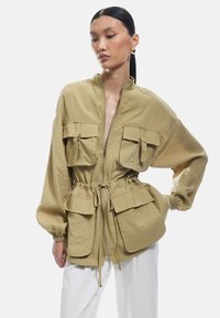Jasnooliwkowa kurtka z zamkiem błyskawicznym, elastycznymi mankietami, dwiema przednimi kieszeniami i sznurkiem w talii. Wykonana z teksturowanej, lekkiej tkaniny.
