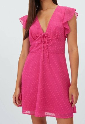 Woman wearing a short pink dress with flutter sleeves, textured fabric, deep V-neck, and tie detail at the chest.