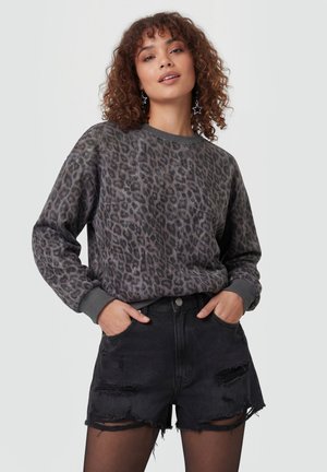 Vrouw met krullend haar die stervormige oorbellen draagt, een grijze leopardprint sweater, zwarte gescheurde spijkershorts en zwarte panty's, handen in de zakken.