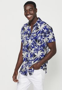Camicetta a maniche corte con sfondo blu e motivi di palme bianche e nere. Design con bottoni, taschino sul lato sinistro del petto.
