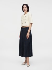 Femme portant une veste courte crème à manches courtes avec des boutons dorés, une jupe large taille haute bleu marine et des chaussures pointues crème.