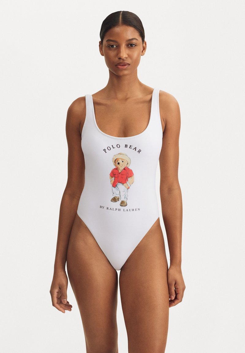 Costumul de baie dintr-o singură piesă este alb și are un grafic cu un urs care poartă o cămașă roșie, cu textul "POLO BEAR" și "BY RALPH LAUREN" imprimat dedesubt.