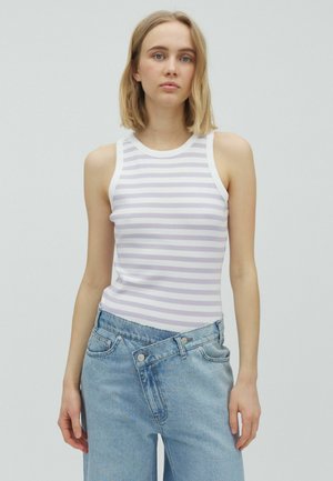 Tânără cu păr blond până la umeri, purtând un top fără mâneci alb cu dungi violet deschis și blugi albaștri cu talie înaltă, pe un fundal gri.