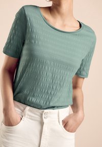 Turkusowa koszulka z krótkim rękawem o teksturowanym wzorze; okrągły dekolt, noszona z białą spódnicą z wysokim stanem, ręce w kieszeniach.