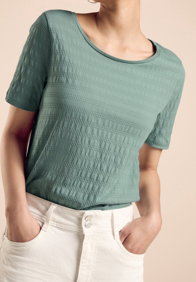 Turkusowa koszulka z krótkim rękawem o teksturowanym wzorze; okrągły dekolt, noszona z białą spódnicą z wysokim stanem, ręce w kieszeniach.