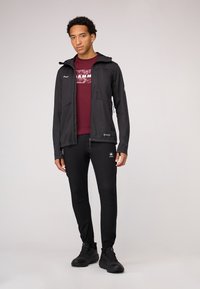 Giacca softshell nera con cappuccio, abbinata a una maglietta color bordeaux e pantaloni neri slim-fit. Include cerniere e polsini elastici. Calzature nere.