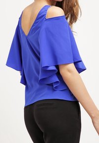 Blå kortärmad toppen med V-formad rygg, volangärmar och dekorerade axeldetaljer, gjord av lätt tyg. Matchad med svarta byxor.