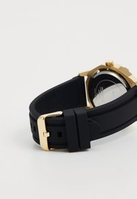 Bracelet de montre en caoutchouc noir avec boucle en or attachée à un boîtier en or, incliné pour montrer les détails de l'arrière et du fermoir sur un fond blanc.