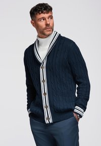 Cardigan a coste navale con colletto a scialle, dettagli a strisce bianche su polsini e patta, dotato di bottoni in legno e tasche laterali.