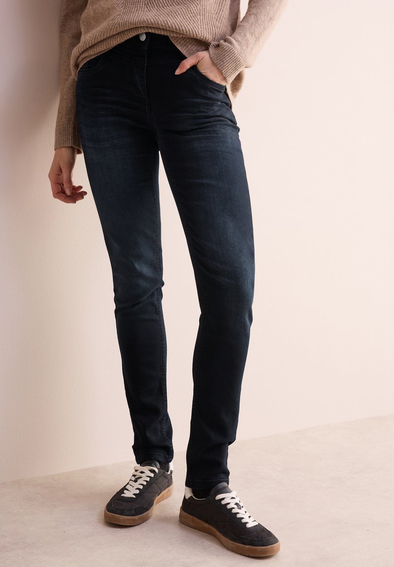 Jeans skinny scuri in denim con una leggera lucentezza, dotati di tasche frontali e vita media, abbinati a sneakers grigie e a un maglione beige.