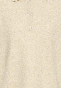Gros plan sur une chemise beige à col avec trois boutons décoratifs nacrés sur la patte, montrant un tissu à la texture douce.