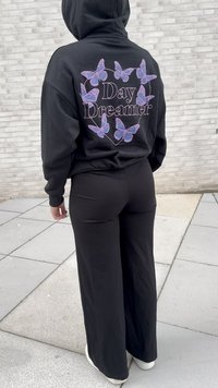 Persoon in een zwarte hoodie en broek, de achterkant van de hoodie toont paarse vlinders die rondom de tekst "Day Dreamer" cirkelen, staand op betegeld trottoir.