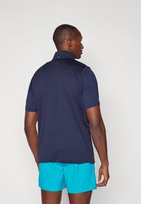 Marineblå kortermet polo med ståkrage, utstyrt med teksturert stoff. Kombinert med lyseblå shorts, vist fra baksiden.