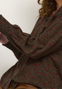 Person wearing a brown long-sleeve shirt with small red flowers and dark leaf patterns, adjusting the sleeve cuff.