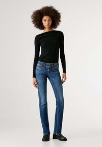 Melns top ar garām piedurknēm, savienots ar ziliem taisna piegriezuma džinsiem ar pogātas jostasvietas un smalkām šuvju detaļām; modelis stāv neitrālā fonā.