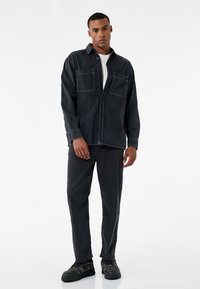 Schwarzes Denim-Hemd mit zwei Fronttaschen, Knopfverschluss und kontrastierenden weißen Nähten, kombiniert mit schwarzen Hosen und strukturierten Sneakers.