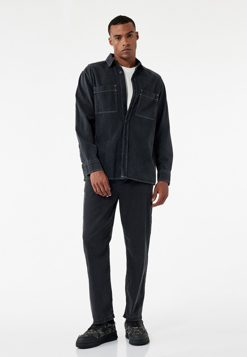 Schwarzes Denim-Hemd mit zwei Fronttaschen, Knopfverschluss und kontrastierenden weißen Nähten, kombiniert mit schwarzen Hosen und strukturierten Sneakers.
