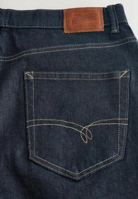 Tmavě modré džínové kalhoty s obdélníkovým zadním kapsou se žlutým šitím a koženou nášivkou s nápisem "DENIM GOODS" nad pasem.