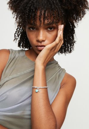 Pulsera de plata con colgante turquesa, con un diseño texturizado. Modelo con un top gris translúcido, sosteniendo su rostro con una mano.