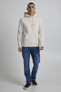 Cremefarvet hoodie med snore og kængurulomme, parret med blå denimjeans og sorte sneakers. Simpelt design, blød tekstur.