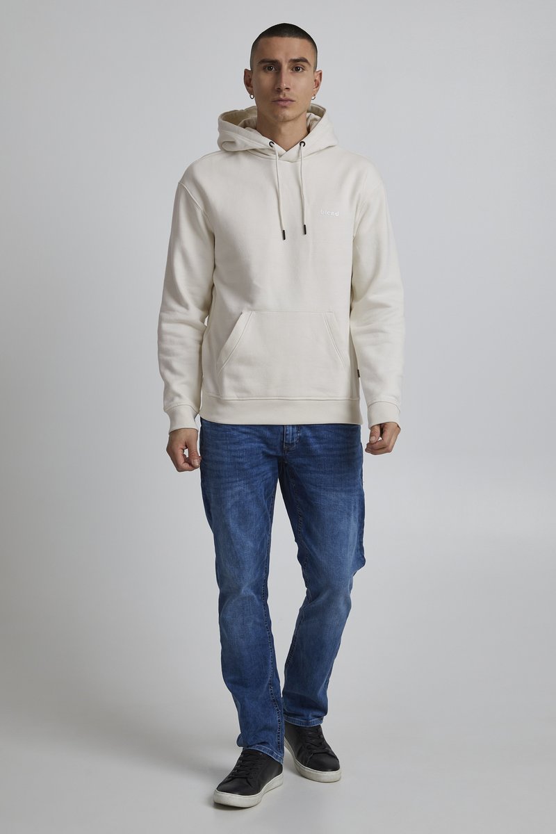 Cremefarvet hoodie med snore og kængurulomme, parret med blå denimjeans og sorte sneakers. Simpelt design, blød tekstur.
