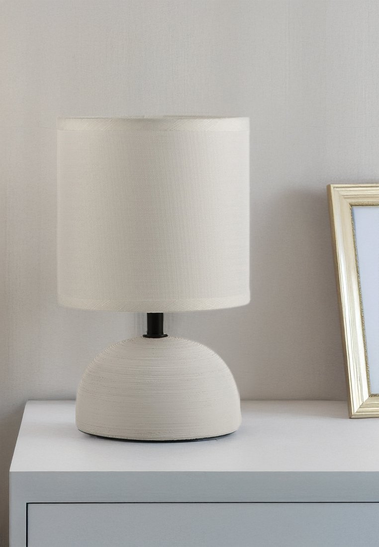 Bílá keramická stolní lampa s látkovým stínidlem na bílém nočním stolku vedle nakloněného fotorámečku se zlatým rámem.