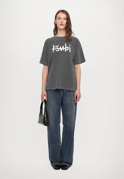 Tricou supradimensionat gri cu logo alb "ksubi", asortat cu blugi evazați bleu. Geantă de mână neagră cu un finisaj texturat și strălucitor. Pantofi negri.