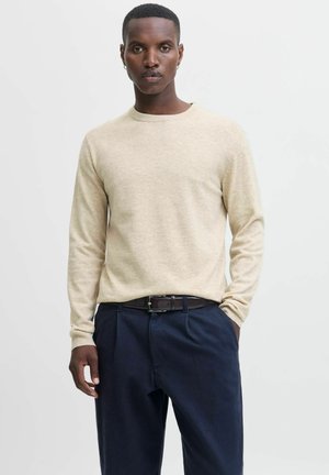 Beige lange mouw trui met een ronde halslijn en een zachte textuur. G gedragen met donkerblauwe broek en een bruine leren riem met een zilveren gesp.