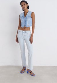 Kratka svetlo modra jeans vesta s sprednjimi gumbi, v kombinaciji s svetlo modrimi flared jeans hlačami in modrimi ter črnimi natikači na peto.