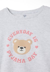 Graues Baumwoll-T-Shirt mit einem Bärenmotiv, der ein hellbraunes Gesicht hat, umgeben von pinkem Text mit der Aufschrift "JEDER TAG IST PYJAMATAG."