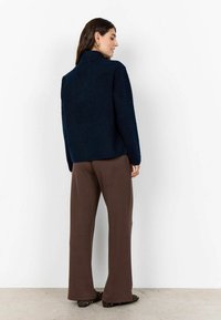 Donkerblauwe gestructureerde jas met een hoge halslijn, gecombineerd met bruine wijde broek en zwarte schoenen met gouden studs.
