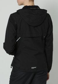 Persona che indossa una giacca con cappuccio Windstopper nera, vista da dietro, su uno sfondo grigio chiaro uniforme.