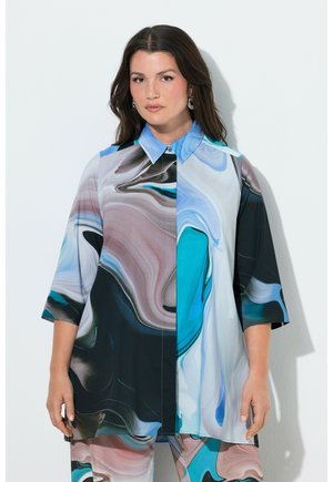 Kobieta ubrana w koszulę na guziki z długim rękawem oraz spodnie z abstrakcyjnym wzorem wirujących motywów w odcieniach niebieskiego, czarnego, białego i różowego.