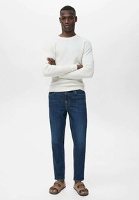 Weiße gerippte Langarmshirt, blaue Jeans, braune Sandalen. Das Modell steht mit verschränkten Armen vor einem schlichten weißen Hintergrund.