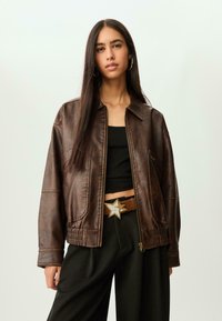 Jeune femme aux longs cheveux bruns portant une veste en cuir marron, un crop top noir, un pantalon large noir et une ceinture avec une boucle en forme d'étoile.
