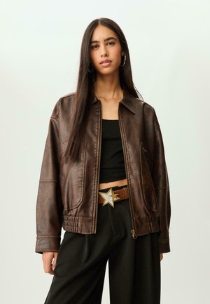 Junge Frau mit langen dunklen Haaren, die eine braune Lederjacke, ein schwarzes Crop-Top, weite schwarze Hosen und einen Gürtel mit einer Sternschnalle trägt.
