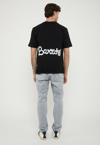 Man with tattoo on left arm wearing black T-shirt with "Barrow" text on back, gray pants, and white sneakers, standing facing away.