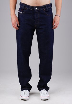 Dunkelblaue Jeans mit geradem Schnitt, ausgestattet mit kupferfarbenen Knöpfen, fünf Taschen und gelben Nähten, kombiniert mit weißen Sneakers.