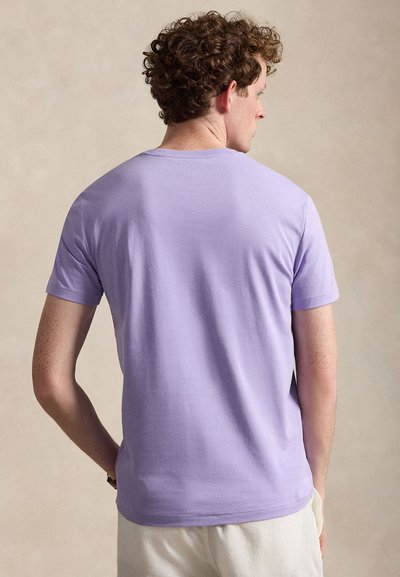 Göndör hajú férfi, aki egyszínű levendula rövid ujjú pólót visel, hátulról látható, semleges háttér előtt.