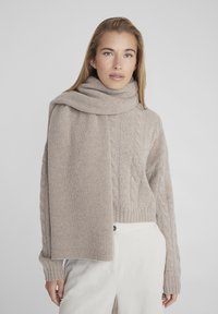 Maglione beige lavorato a maglia con trama a trecce e coste, abbinato a una sciarpa coordinata. Tessuto morbido, vestibilità rilassata. Il modello indossa pantaloni di colore chiaro.